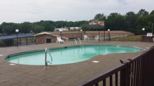 outdoor pool with paved surround, pool lounger and chairs, surrounded by fencing and lights