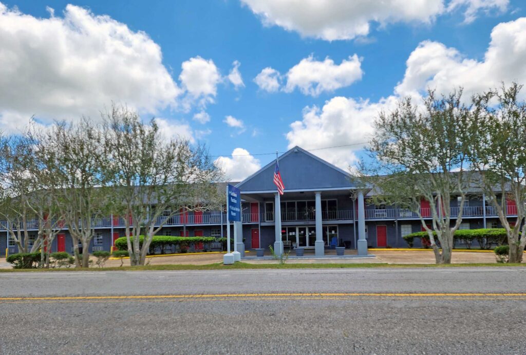 Two story building with canopied drive through entrance, exterior guest room entrances with covered walkway, grassy area with trees, American flag pole