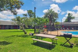 outdoor pool with fencing, patio tables with umbrella and chairs, sun loungers, one story hotel building overlooking pool with exterior guest room entrances, grassy area and trees, guest BBQ area and picnic table with benches, mermaid statue