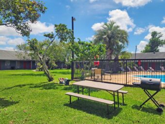 outdoor pool with fencing, patio tables with umbrella and chairs, sun loungers, one story hotel building overlooking pool with exterior guest room entrances, grassy area and trees, guest BBQ area and picnic table with benches, mermaid statue