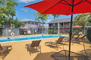 outdoor pool with fencing, patio tables with umbrella and chairs, sun loungers, two story hotel building overlooking pool with exterior guest room entrances