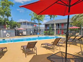 outdoor pool with fencing, patio tables with umbrella and chairs, sun loungers, two story hotel building overlooking pool with exterior guest room entrances