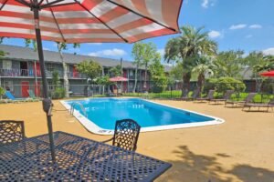 outdoor pool with fencing, patio tables with umbrella and chairs, sun loungers, two story hotel building overlooking pool with exterior guest room entrances