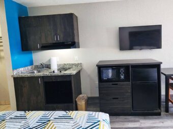 Wall cabinets with cooker hood, floor cabinet with sink and hob, wooden unit with fridge and microwave, wall mounted TV, table and chair, laminate flooring